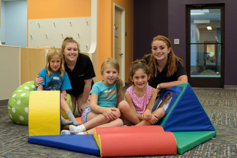 Two adults and three children sitting on a carpeted floor in a colorful indoor play area, surrounded by soft foam play shapes in bright colors like yellow, red, blue, and green.