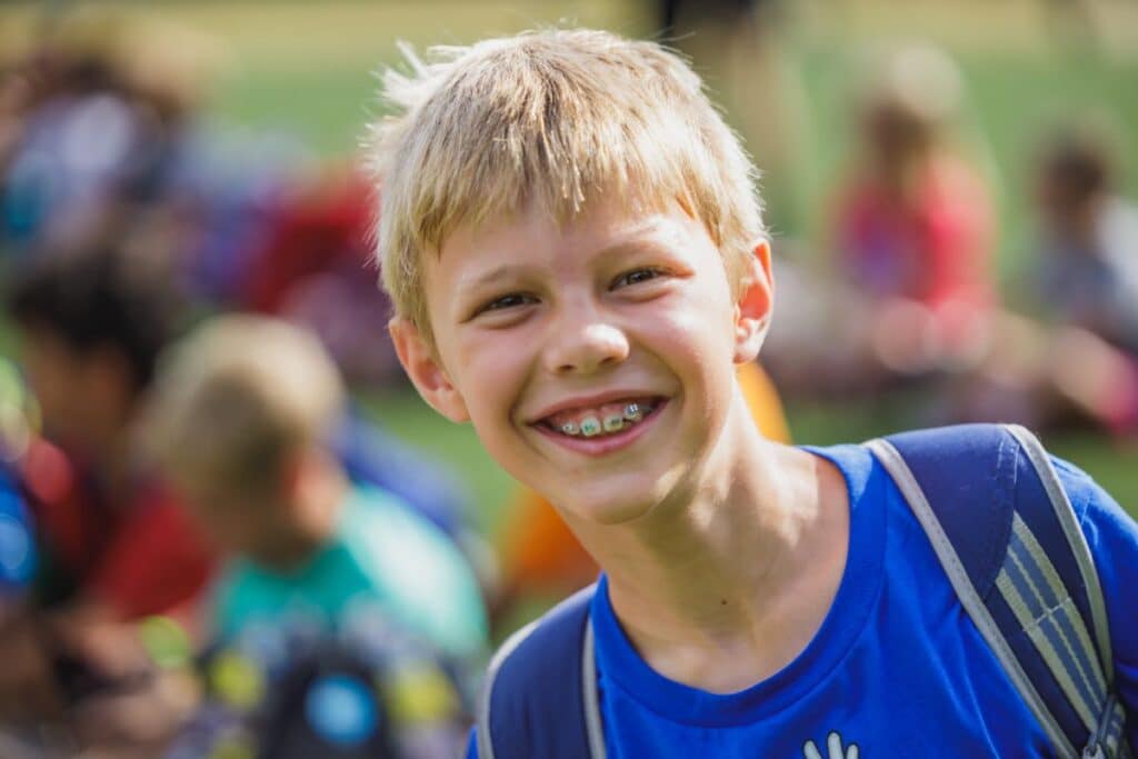 boy with blonde hair and braces smiling at camp with other campers behind him