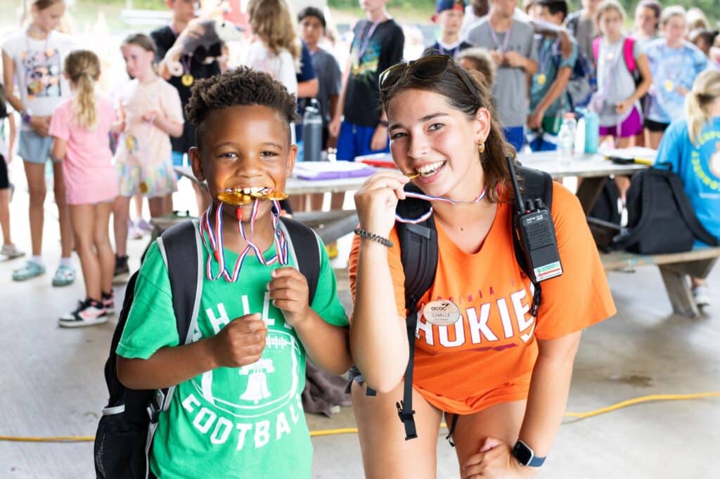 camper in a green shirt and counselor in an orange shirt biting gold medals during olympic themed camp week