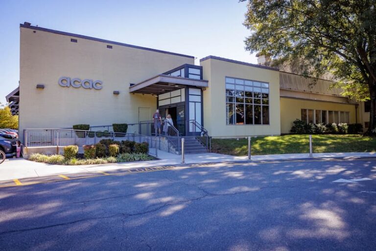 The image shows the exterior of an ACAC Fitness & Wellness Center building. It has a light yellow facade with the ACAC logo displayed on the left side. The entrance features a modern design with large glass windows, a metal awning, and a ramp with railings leading up to the doors. There is landscaping with shrubs along the front and a tree providing shade on the right side. The setting appears to be a sunny day with clear skies.