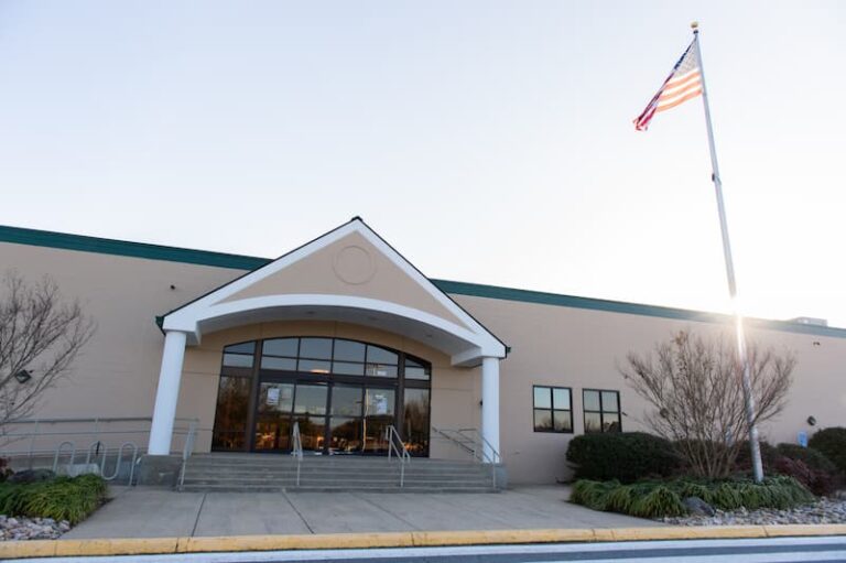 The image shows the entrance of a large beige building with a green roof trim and a prominent arched design above the doorway. There are wide glass doors and windows at the front, flanked by two white columns. A set of steps leads up to the entrance, with metal railings on both sides. Landscaping with shrubs and plants surrounds the area, and an American flag is mounted on a tall flagpole to the right. The sky is clear, and the lighting suggests late afternoon or early evening.