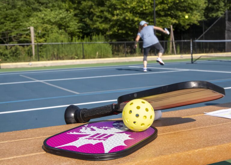 CopilotPickleball paddles and a yellow perforated ball resting on a wooden bench in the foreground, with an outdoor pickleball court in the background where a person is actively playing. The court has blue playing surfaces, white boundary lines, and is surrounded by a black chain-link fence with green trees beyond.