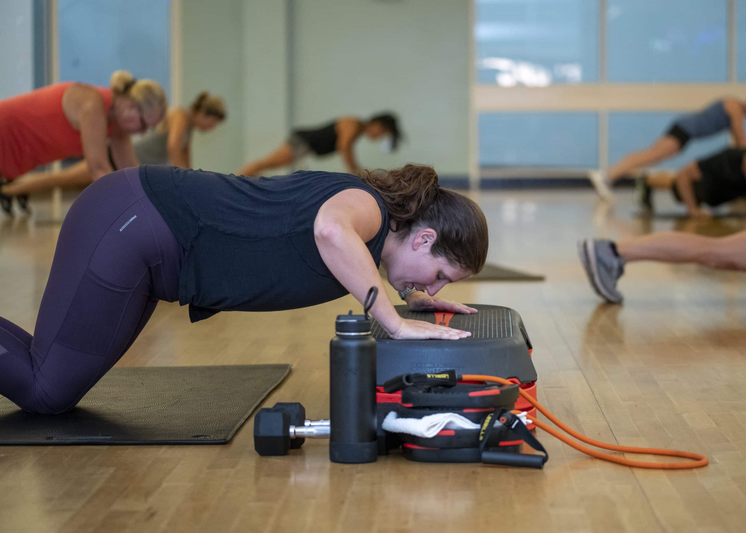 Person performing a modified push-up on a step platform in a fitness studio, with resistance bands, a dumbbell, and a water bottle nearby. Other participants are exercising in the background.