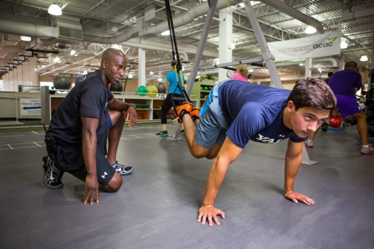 Person performing a suspension training exercise in a gym, with feet strapped into suspension straps and body in a plank position, while another person kneels nearby observing. The background shows gym equipment, exercise balls, and other people working out.