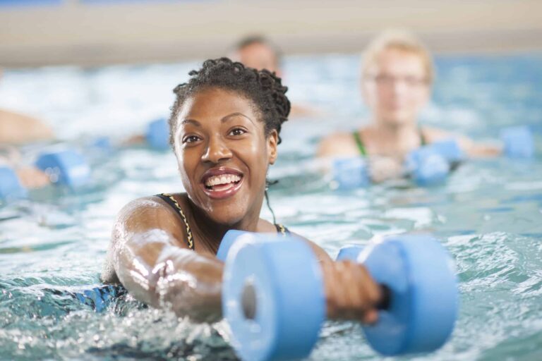 Person participating in an aquatic fitness class, holding two blue foam dumbbells while standing in a swimming pool. Other participants with similar equipment are visible in the background, and the water is clear with gentle ripples. The setting suggests a group water exercise session focused on strength and resistance training.