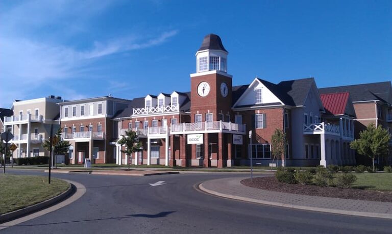 A large brick building with a clock tower and white trim, featuring the ACAC logo above the entrance. The structure has multiple sections with peaked roofs and balconies, set along a circular driveway with landscaped greenery. The sky is clear and blue, indicating a sunny day.