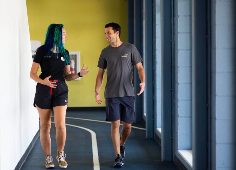 Two people walking and talking on an indoor track with blue lanes and white markings, next to large windows and a yellow wall in the background. Both are wearing athletic clothing and sneakers.