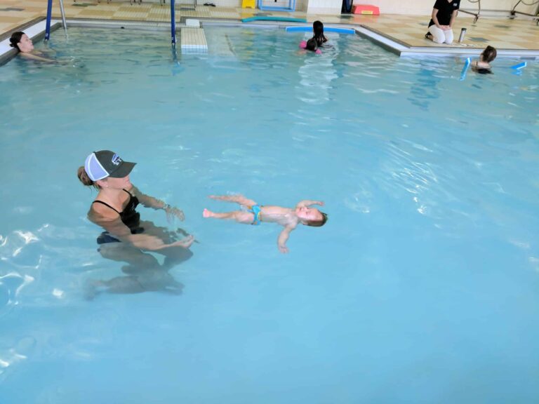 Indoor swimming pool with a baby floating on its back in the water while an adult wearing a black swimsuit and a cap is positioned nearby for support. Other people are visible in the background, some sitting at the pool’s edge and others in the water. The pool area has tiled flooring and walls, with swim equipment placed around the perimeter.