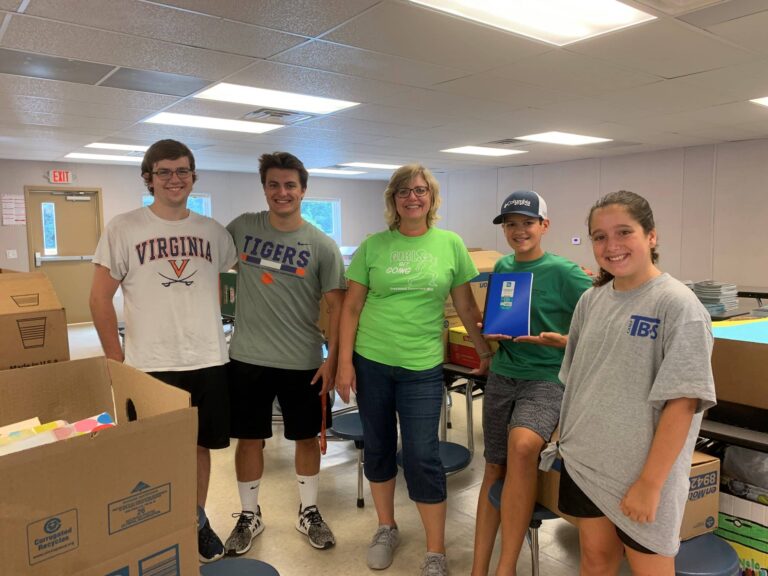 A group of five people standing together in a room with tables and large cardboard boxes filled with supplies. One person is holding a blue notebook, and others are wearing casual clothing, including T-shirts with text such as “Virginia” and “Tigers.” The setting appears to be a community center or classroom, with fluorescent lighting, a tiled ceiling, and windows in the background. The scene suggests an activity related to organizing or distributing school supplies