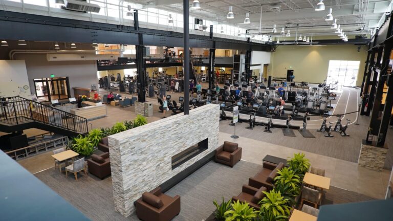 Spacious modern fitness center with an open layout featuring multiple workout areas. The foreground includes a lounge area with cushioned chairs, small tables, and decorative plants surrounding a large stone fireplace. In the background, there are rows of cardio machines such as treadmills, ellipticals, and stationary bikes, along with strength training equipment. The gym has high ceilings with exposed beams, bright lighting, and large windows providing natural light. A staircase leads to an upper level with additional exercise equipment.