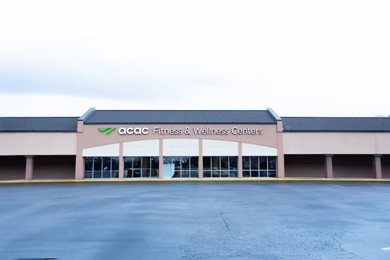 The image shows the exterior of an ACAC Fitness & Wellness Centers location. The building has a beige facade with a dark roof and large glass windows at the entrance. The ACAC logo and text are prominently displayed above the doorway. The foreground features a wide paved area, likely a parking lot, and the sky appears overcast.