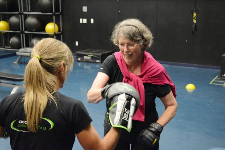 Two people are in a gym setting engaged in a boxing training session. One person is wearing black gloves and a pink sweatshirt draped over their shoulders, throwing a punch toward the other person, who is holding padded boxing mitts. The background includes gym equipment such as large exercise balls stacked on shelves, resistance bands, and a blue floor with yellow markings. The scene suggests a personal training or fitness activity focused on boxing techniques.