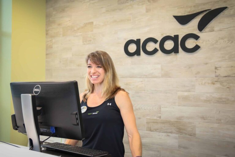 Reception area with a light wood wall featuring the ACAC logo in black lettering and a stylized leaf design. A person is standing behind a desk working on a Dell computer, wearing a black athletic tank top with an Under Armour logo and an ACAC name badge. The setting appears clean and modern, with neutral tones and a professional atmosphere.