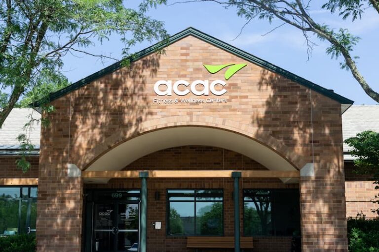 The image shows the entrance of an ACAC Fitness & Wellness Centers building. It features a brick exterior with an arched design above the doorway, the ACAC logo prominently displayed near the top, and two green support columns at the entrance. There is a bench to the right and trees framing the view.