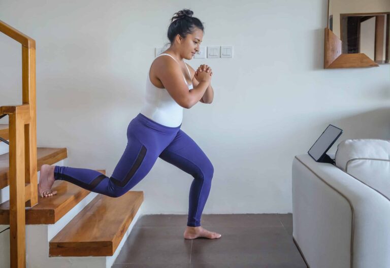 Person performing a Bulgarian split squat exercise indoors. One foot is elevated on a wooden staircase behind, while the other foot is firmly on the floor in a lunge position. The individual is wearing a white tank top and purple leggings, with hands clasped together in front of the chest. A tablet is positioned on a white couch nearby, likely for following a workout routine. The setting appears to be a home environment with neutral-colored walls and tiled flooring.
