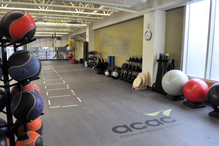 Spacious gym area with gray flooring featuring the ACAC Fitness & Wellness Center logo, lined with various exercise equipment including medicine balls on a rack, kettlebells, dumbbells, resistance bands, and large stability balls. The room has high ceilings with exposed beams, large windows allowing natural light, and a wall clock visible on the right side.