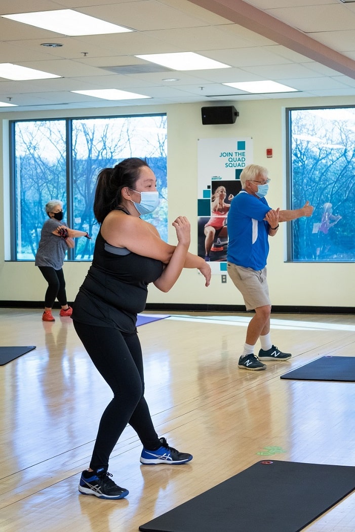 People participating in a fitness class in a bright studio with large windows. They are standing on exercise mats and performing a stretching or warm-up movement with arms extended across the body. The room has wooden floors, wall-mounted speakers, and a poster that reads “JOIN THE QUAD SQUAD.”