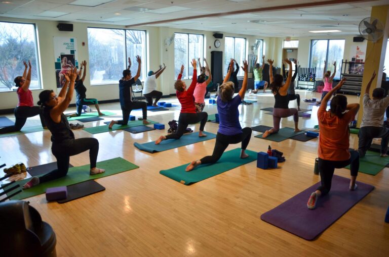 A group of people participating in a yoga class in a spacious studio with wooden floors and large windows. They are positioned on yoga mats in a lunge pose with arms raised overhead. Yoga blocks and other props are visible on the floor, and the room is well-lit with natural light coming through the windows.