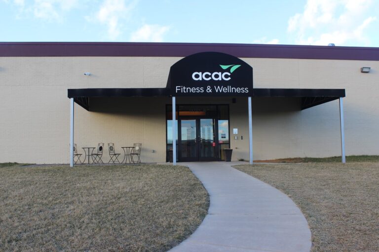 Entrance of a fitness and wellness center with a black awning displaying the logo and text “acac Fitness & Wellness.” The building has light-colored walls, a concrete walkway leading to the glass doors, and a small outdoor seating area with metal tables and chairs on the left side.