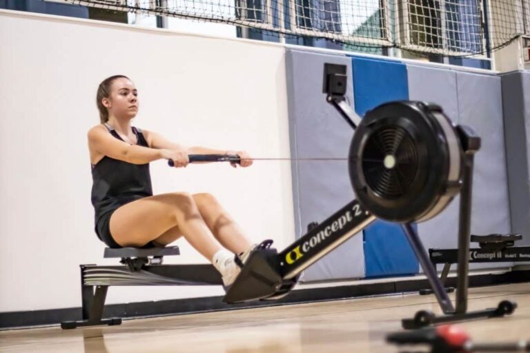Person using a Concept2 rowing machine in an indoor gym, pulling the handle while seated with legs extended. The background includes padded wall panels and a netted area above.