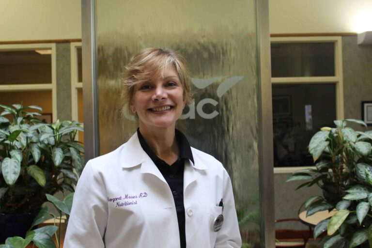 Person wearing a white lab coat standing indoors near a glass panel with the letters “AC” partially visible, surrounded by green plants. The lab coat has embroidered text on the left side and a pen in the pocket.