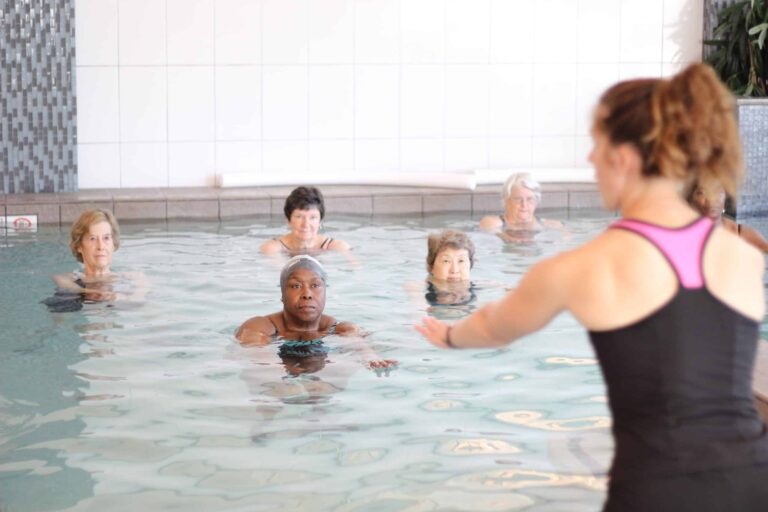 Indoor pool setting with an instructor standing at the edge of the water, guiding a group of participants who are partially submerged in the pool. The participants are spaced out in a line facing the instructor, and the background features white tiled walls and a section of mosaic tiles. The water is clear, and the scene suggests an aquatic fitness class or swimming lesson.