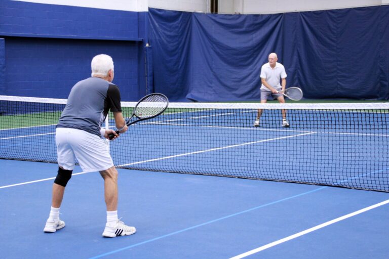 Two older adults playing tennis on an indoor court with blue flooring and a net in the center.