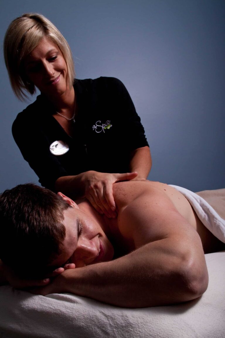 Person lying face down on a massage table with a white towel covering the lower body, receiving a shoulder and upper back massage from a massage therapist wearing a black top with a spa logo. The background is a plain blue wall, creating a calm and professional setting.