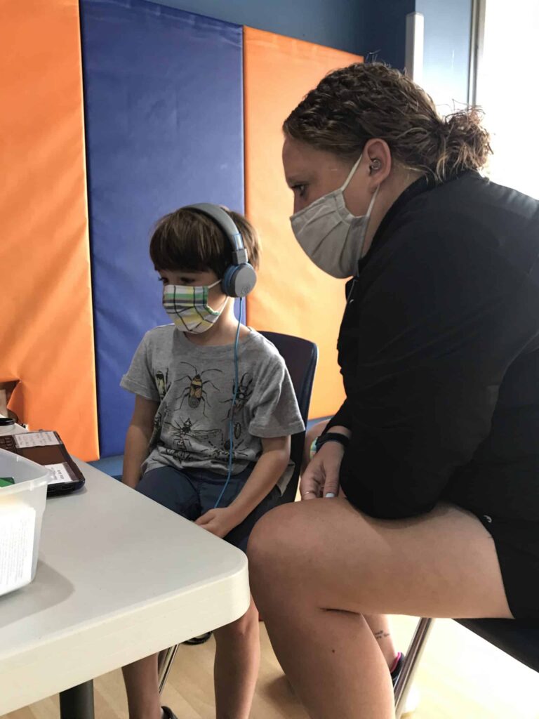 A child wearing headphones sits at a table with a tablet or electronic device, while an adult sits nearby, leaning in as if assisting or supervising. The background features padded panels in orange and blue colors.