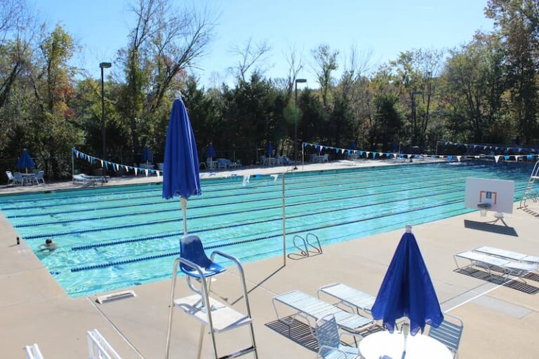 An outdoor swimming pool with multiple lap lanes marked by blue and white lane dividers. The pool deck has lounge chairs, tables, and blue umbrellas arranged around the perimeter. Trees and greenery surround the area, and the sky is clear and sunny, indicating a pleasant day for swimming.