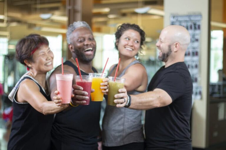 Four people standing together inside a fitness center holding large plastic cups filled with colorful smoothies. The drinks include pink, red, yellow, and green varieties, each with a red straw. The background shows gym equipment and exercise posters, indicating the setting is a workout or wellness environment.