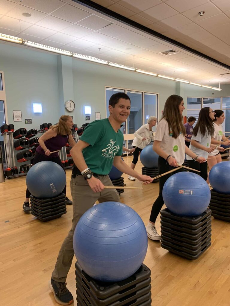 Group fitness class in a studio with wooden floors, where participants are performing a drumming-style workout using drumsticks and large blue exercise balls placed on top of stacked platforms. The room has light-colored walls, mirrors, and racks of weights in the background, with bright overhead lighting.