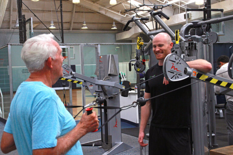 Two people in a gym setting using a cable machine. One person is actively performing a cable exercise, pulling the handles outward, while the other stands nearby, appearing to provide guidance or instruction. The background includes additional fitness equipment, such as weight machines and a basketball court enclosed by netting. The gym has high ceilings with exposed beams and bright overhead lighting.