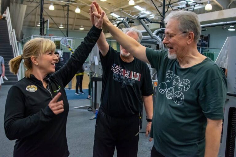 Three people standing in a gym near exercise machines, raising their arms together in a high-five gesture. The setting includes fitness equipment, a staircase, and bright overhead lighting.
