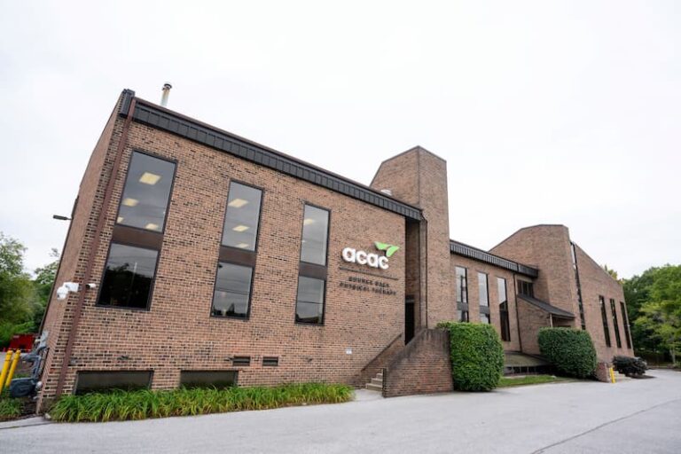 A brick building with large rectangular windows and the ACAC logo on the front, along with the words “Fitness & Wellness.” The structure has a modern design with clean lines, landscaped greenery along the base, and a paved driveway in front. Trees are visible in the background under an overcast sky.