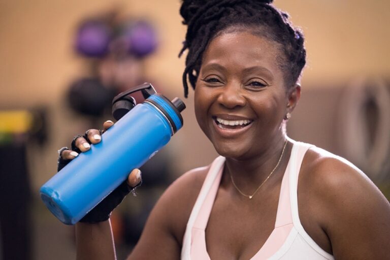 Person in athletic wear holding a blue water bottle in a gym setting, with exercise equipment visible in the background.