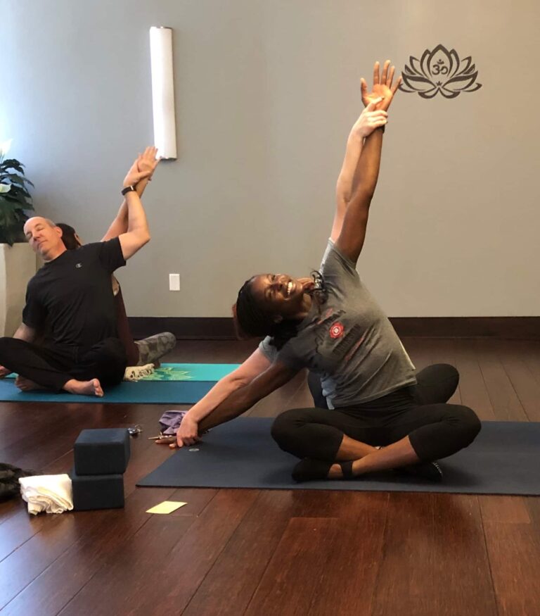 Two people seated on yoga mats in a studio with wooden floors, performing a side stretch with one arm extended upward and the other reaching toward the floor. Yoga blocks and a towel are visible nearby, and a lotus design decorates the wall in the background.