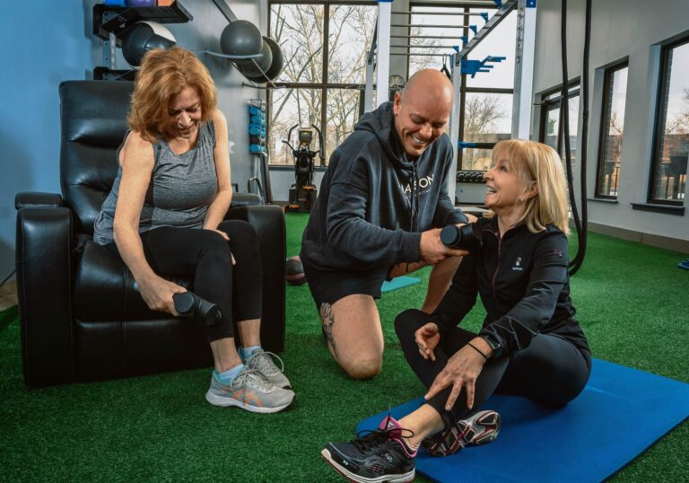 Three people in a gym setting, with two seated on the floor and one in a chair, holding small dumbbells during a workout session. Exercise equipment and large windows are visible in the background.