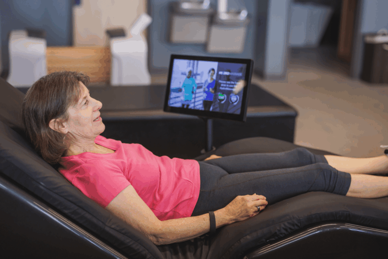 Person reclining on a black lounge chair in a relaxation area, wearing a pink top and dark pants. A screen in front displays fitness-related content, and water fountains are visible in the background.