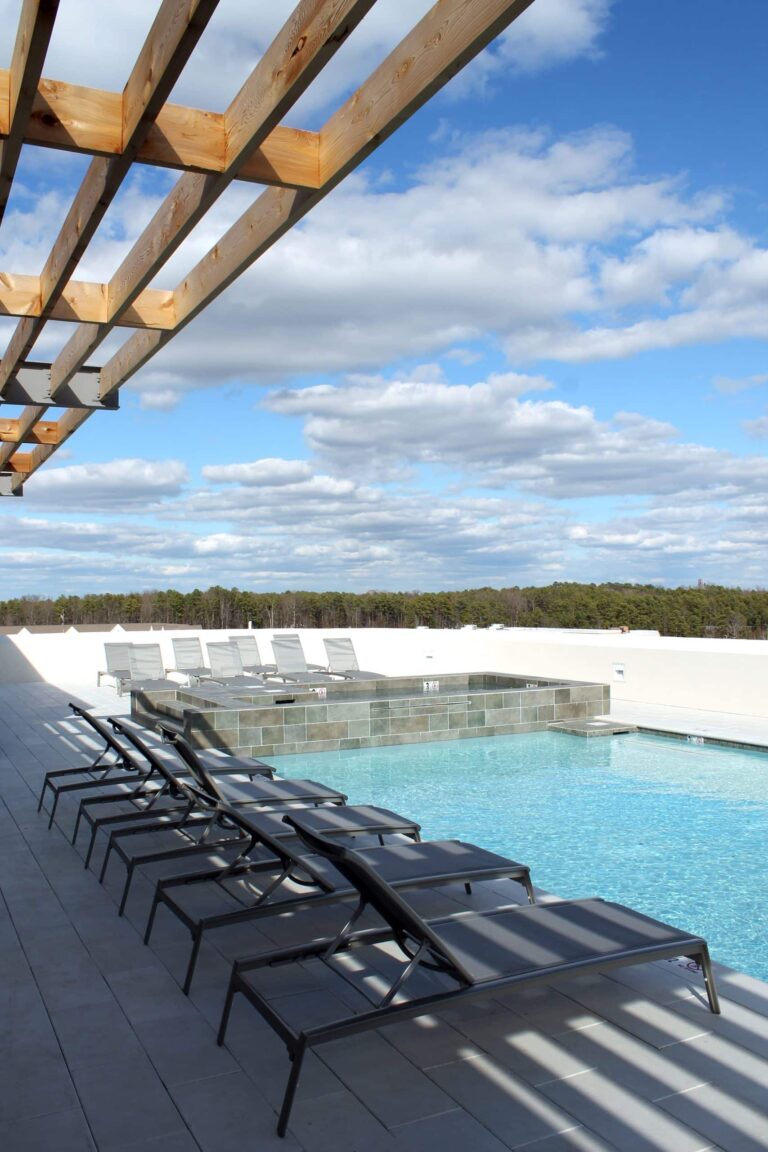 Outdoor pool area with clear blue water, multiple lounge chairs lined up on the deck, and a wooden pergola overhead under a partly cloudy sky.