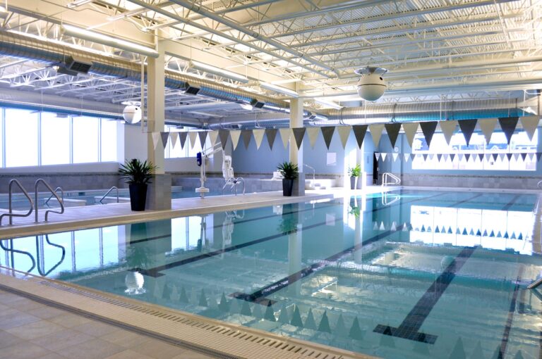 Indoor swimming pool area with clear water, lane markings, and triangular flags hanging above for lap swimming. The space has bright overhead lighting, large windows along one side providing natural light, and potted plants placed near the pool edges. The pool deck is tiled, and there is a smaller adjacent pool visible in the background.