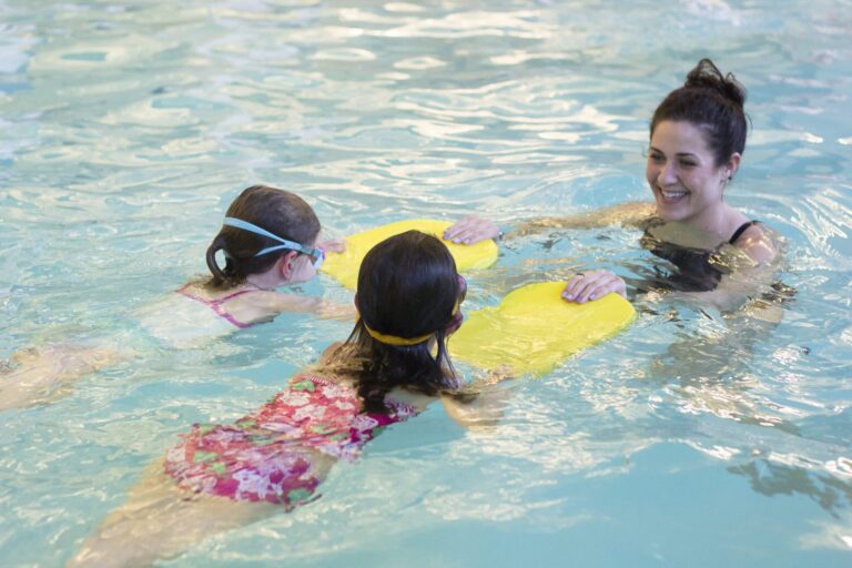 Two children in a swimming pool using bright yellow kickboards while an adult provides guidance. The water is clear, and the setting appears to be an indoor swimming lesson or practice session.