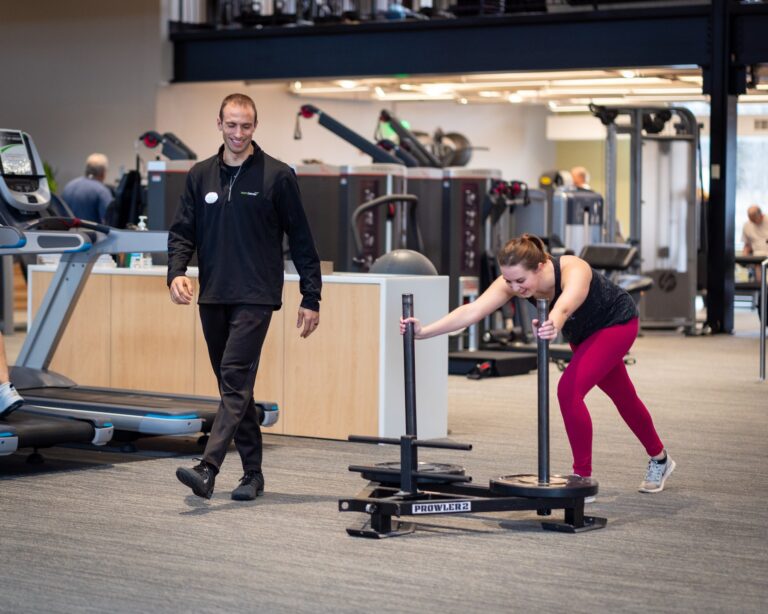 Person pushing a weighted sled labeled “Prowler 2” across a gym floor while another individual walks nearby. The gym setting includes treadmills, strength training machines, and other fitness equipment in the background, with bright overhead lighting and an open layout.