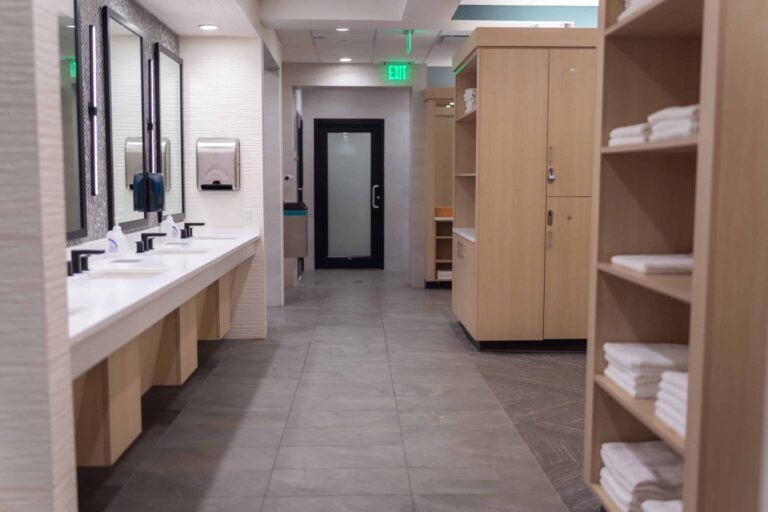 Modern locker room with light wood cabinets and shelves stocked with folded white towels, a row of sinks with mirrors on the left, and a door at the end of the tiled hallway