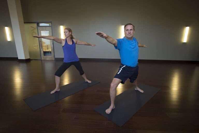 Two people practicing yoga in a studio, standing on mats in a warrior pose with arms extended and legs apart.