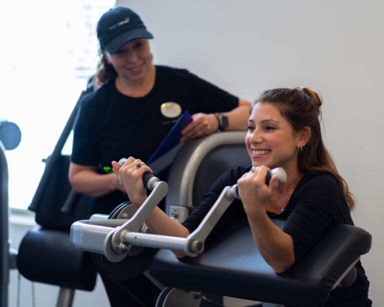 Person using a seated arm curl machine for strength training, gripping the handles with both hands. Another individual stands nearby, holding a clipboard and wearing a dark shirt and cap, likely providing guidance or instruction. The setting appears to be a gym or fitness center with light-colored walls and exercise equipment in the background.