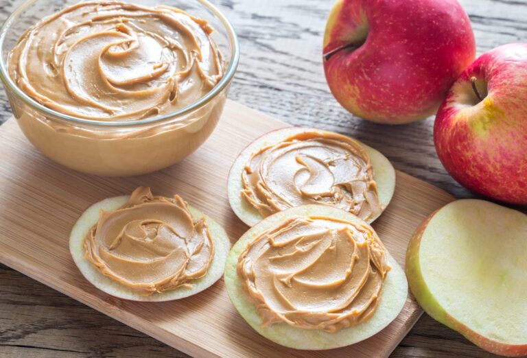 Apple slices spread with creamy peanut butter on a wooden board, next to a glass bowl filled with peanut butter and several whole red apples.
