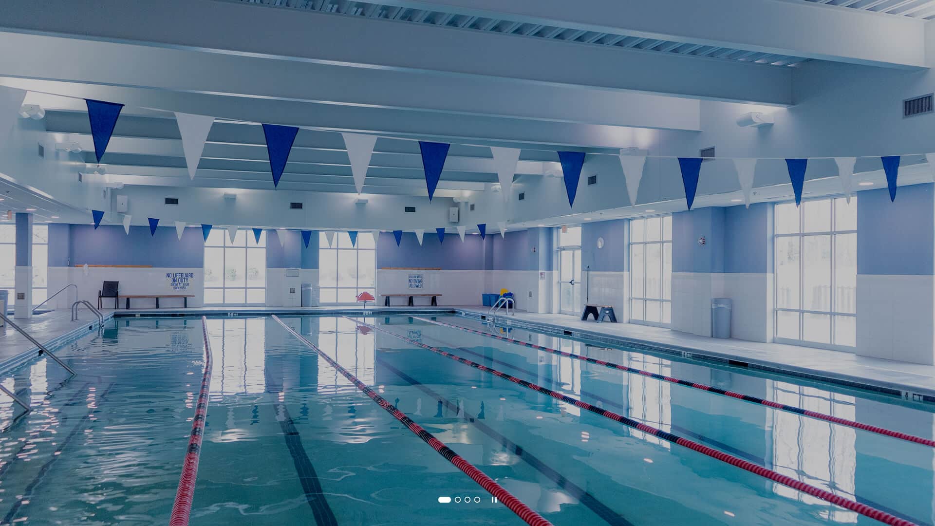 Indoor swimming pool with multiple lanes marked by red lane dividers, blue triangular flags hanging overhead, and large windows allowing natural light into the space. Benches and pool equipment are visible along the walls.