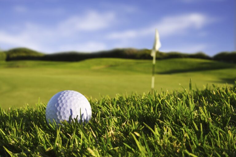 Close-up of a golf ball on green grass with a flagstick and hole in the background under a blue sky.