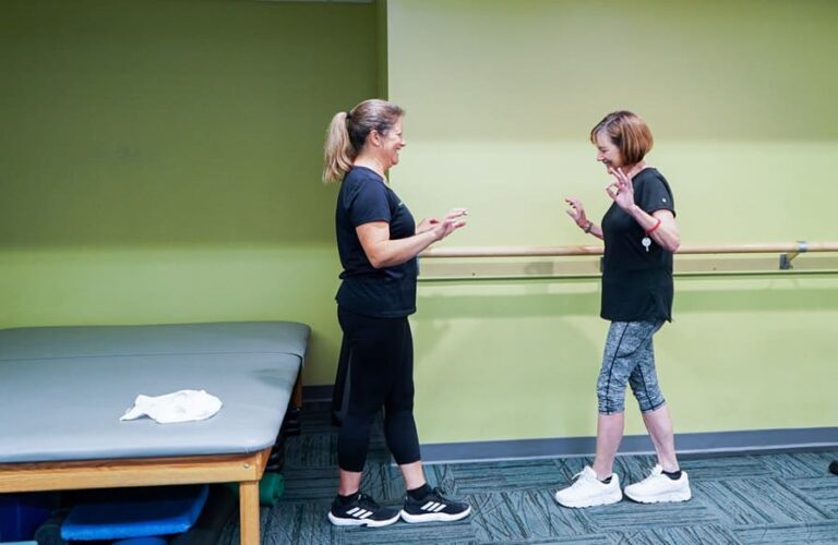 Two people standing in a fitness or physical therapy room, engaged in conversation near a wall-mounted balance bar. One person is wearing athletic shoes and patterned leggings, while the other is in black workout attire. A padded therapy table with a towel is visible to the side, and the room has green walls and carpeted flooring.